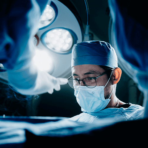 Cirurgião paramentado com máscara, touca e óculos durante procedimento em centro cirúrgico, sob iluminação cirúrgica, visto entre outros profissionais da equipe médica.